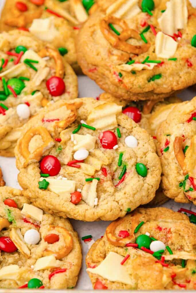 Close-up of chewy Christmas snack cookies packed with red and green M&Ms, white chocolate chunks, crushed potato chips, mini pretzels, and festive sprinkles.