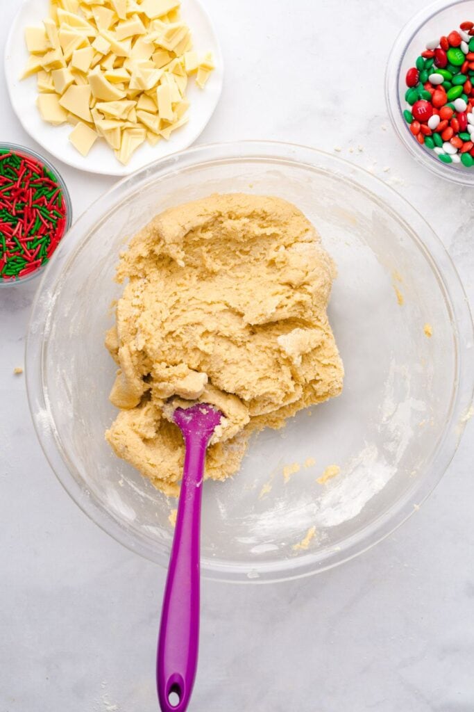 Thick cookie dough in a clear mixing bowl with a purple spatula, surrounded by bowls of red and green holiday sprinkles, chopped white chocolate, and Christmas M&Ms, ready for mix-ins.