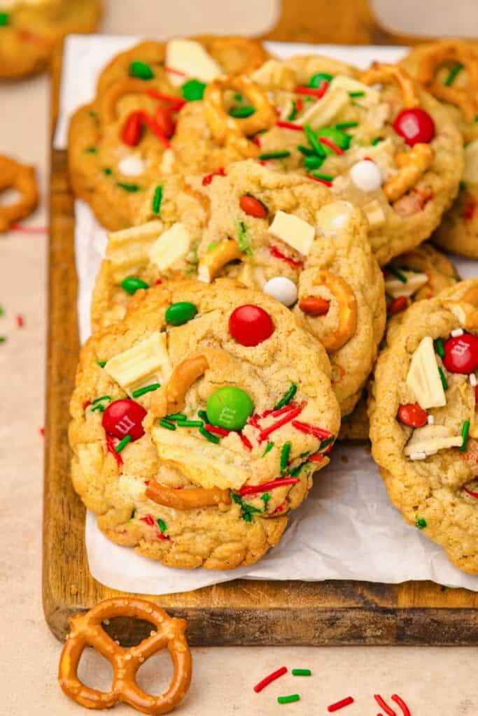 A wooden cutting board with Kitchen Sink Holiday Cookies loaded with red and green candies, white chocolate chunks, crunchy pretzel pieces, and festive red, green, and white sprinkles.