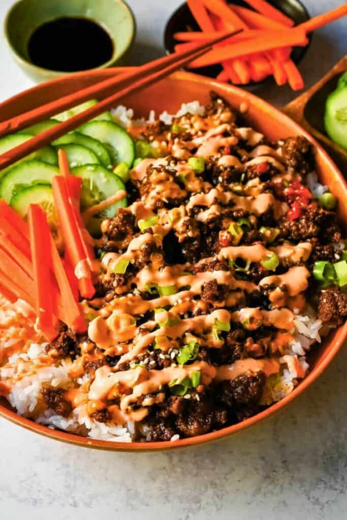 Korean beef bowls with seasoned ground beef served over white rice topped with sliced cucumbers, carrot sticks, green onions, and sesame seeds. The bowl is drizzled with a creamy spicy sauce and served with chopsticks and small side dishes.