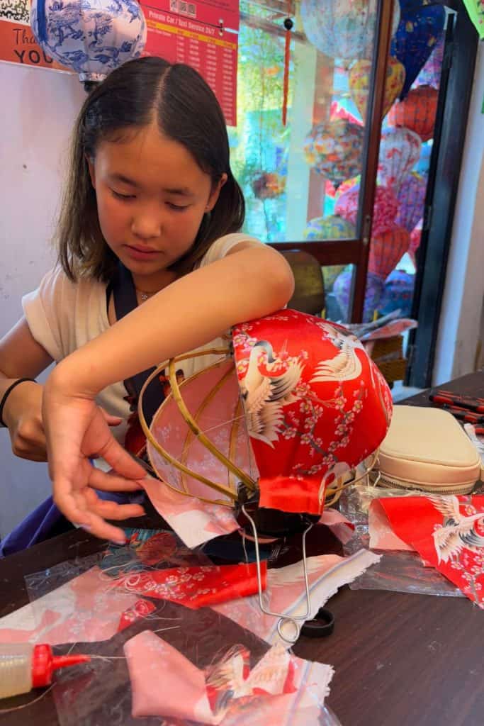 A young girl carefully assembles a traditional Vietnamese silk lantern, wrapping red and pink fabric with crane and cherry blossom patterns around a bamboo frame. She sits at a table surrounded by fabric scraps, thread, and tools, with finished lanterns hanging in the colorful workshop behind her