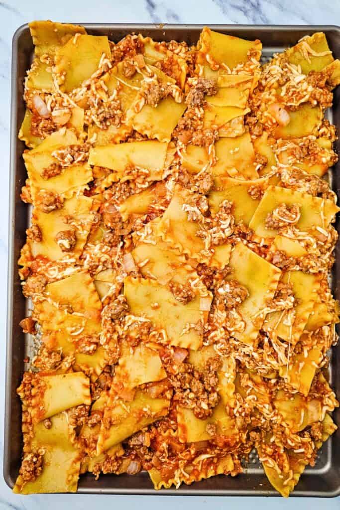 Lasagna noodles mixed with Italian sausage meat sauce and shredded cheese spread across a large sheet pan. The mixture forms an even layer ready for the remaining toppings before baking.