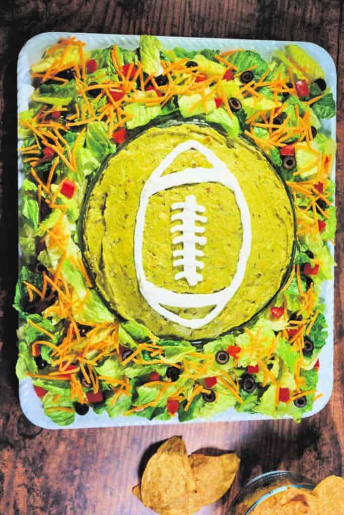 Layered taco dip styled for game day, featuring a large round section of guacamole in the center decorated with sour cream in the shape of a football, surrounded by shredded lettuce, carrots, olives, and diced red peppers, perfect for scooping with chips as a festive game day dip.