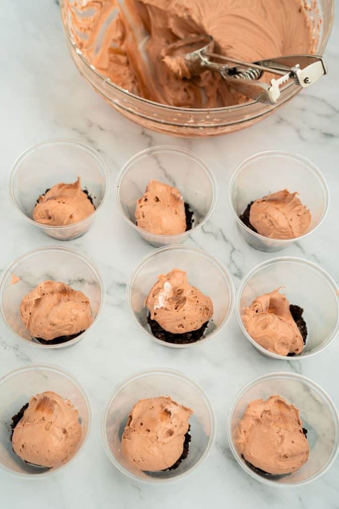 Clear plastic cups partially filled with crushed cookies and topped with scoops of chocolate pudding mixture, with a mixing bowl and cookie scoop in the background, showing the assembly of pudding dirt cups.