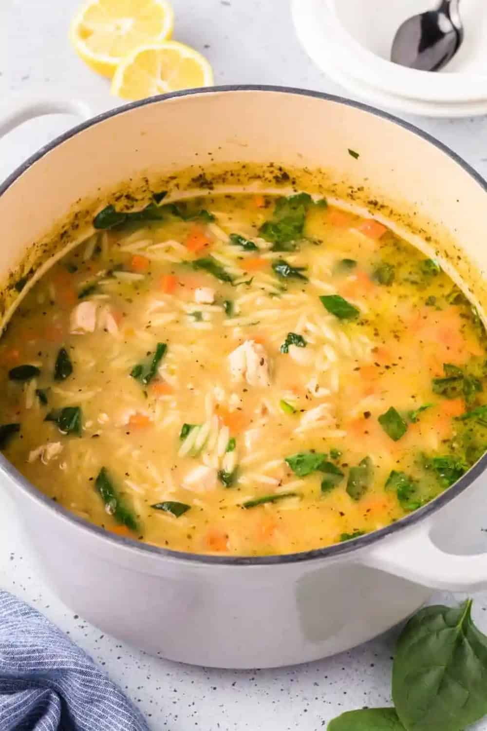 Lemon chicken orzo soup with tender chicken orzo carrots and spinach simmers in a golden lemon broth. The hearty soup is shown in a large white Dutch oven with fresh lemon halves nearby.