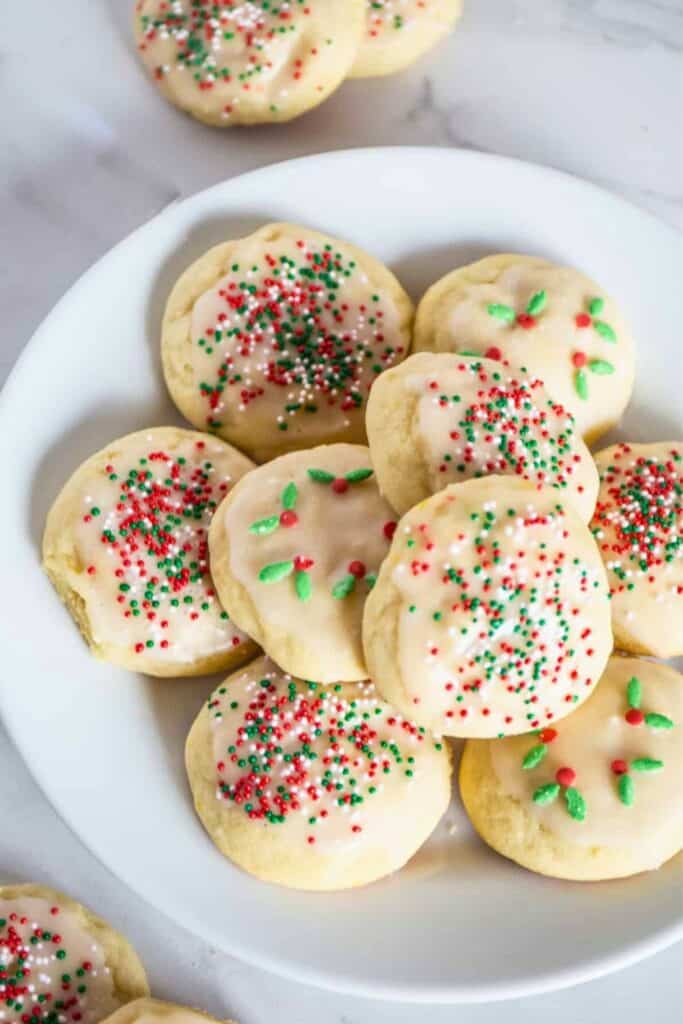 Lemon Italian ricotta cookies are a classic holiday treat topped with a light glaze and festive red, green, and white sprinkles are arranged on a white plate.