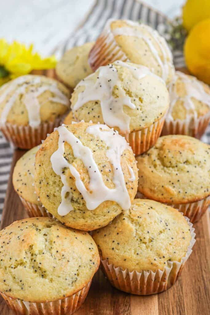 Stack of fluffy lemon poppy seed muffins with a golden crust, some topped with a white citrus glaze drizzle. The muffins are speckled with poppyseeds and arranged on a wooden board, offering a bright, zesty treat perfect for breakfast or brunch.