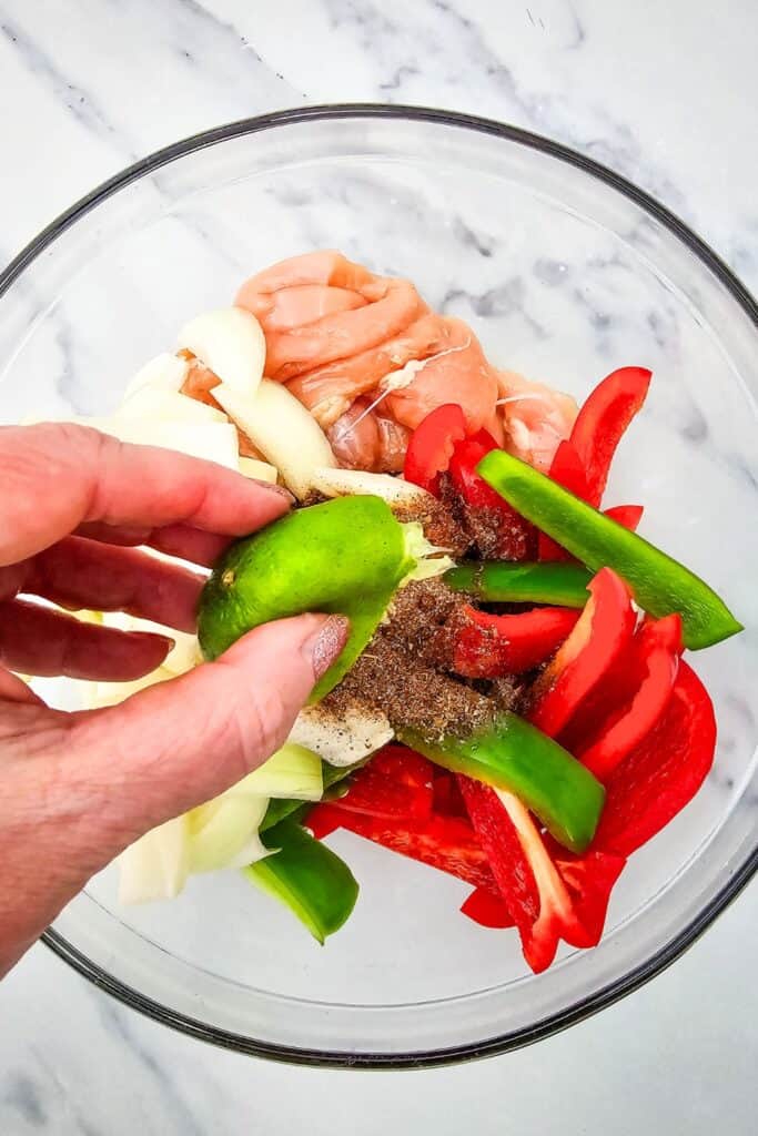Hand squeezing fresh lime juice over raw chicken strips, sliced red and green bell peppers, and onions in a glass bowl with visible fajita seasoning on top.