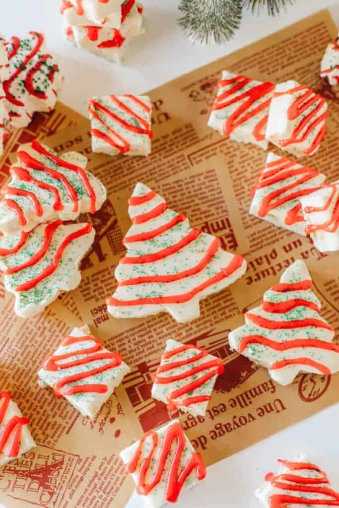 White chocolate Christmas fudge cut into festive shapes, including Christmas trees, decorated with red icing swirls and green sugar sprinkles. The treats are arranged on vintage-style parchment paper, resembling the look of Little Debbie holiday snacks.