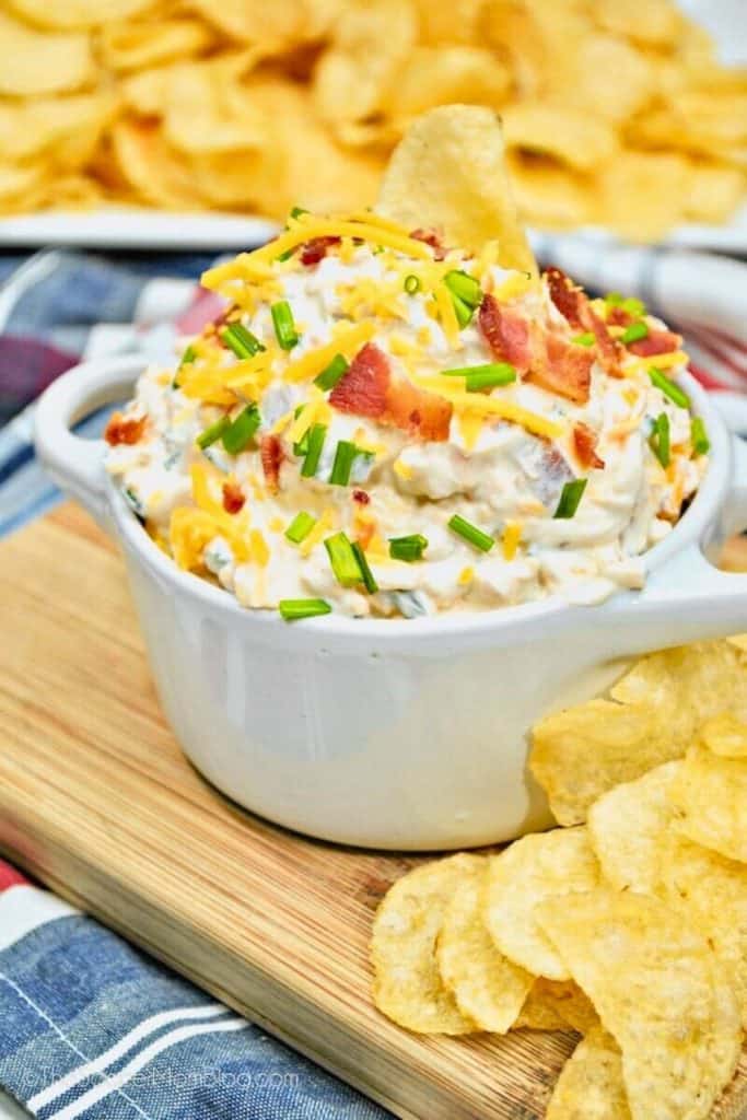Loaded Baked Potato Dip in a white ceramic bowl, topped with shredded cheddar cheese, crispy bacon pieces, and chopped chives. Served with ridged potato chips on a wooden board, capturing all the flavors of a baked potato in a creamy, dippable form.