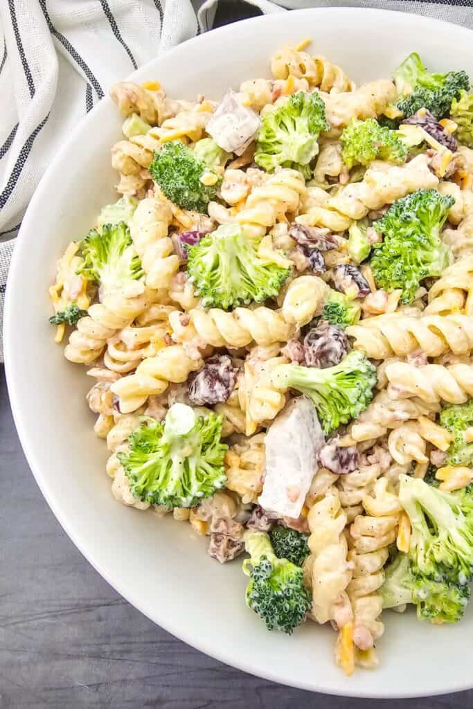 Overhead view of a loaded broccoli pasta salad in a white bowl, packed with rotini pasta, fresh broccoli florets, bacon bits, shredded cheese, dried cranberries, and chunks of chicken, all mixed in a creamy dressing.