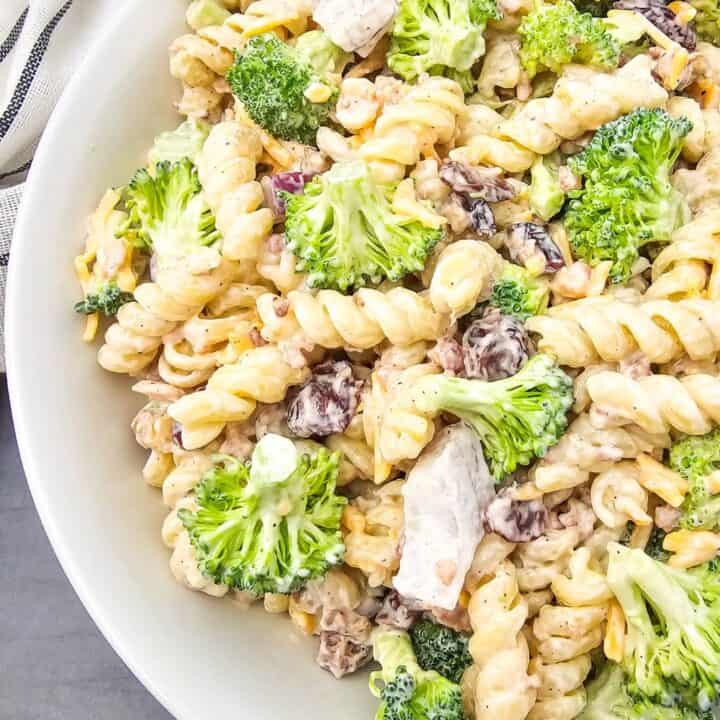 Overhead view of a loaded broccoli pasta salad in a white bowl, packed with rotini pasta, fresh broccoli florets, bacon bits, shredded cheese, dried cranberries, and chunks of chicken, all mixed in a creamy dressing.