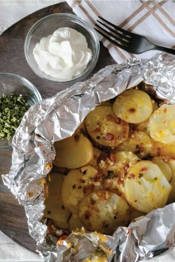 A tin foil pouch of loaded cheesy sliced potatoes beside two small bowls of chives and sour cream. 