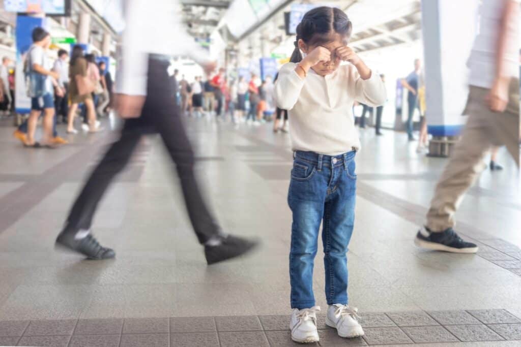 Young child standing alone in a busy transit station rubbing their eyes while blurred crowds walk past. The image illustrates a lost child while travelling.