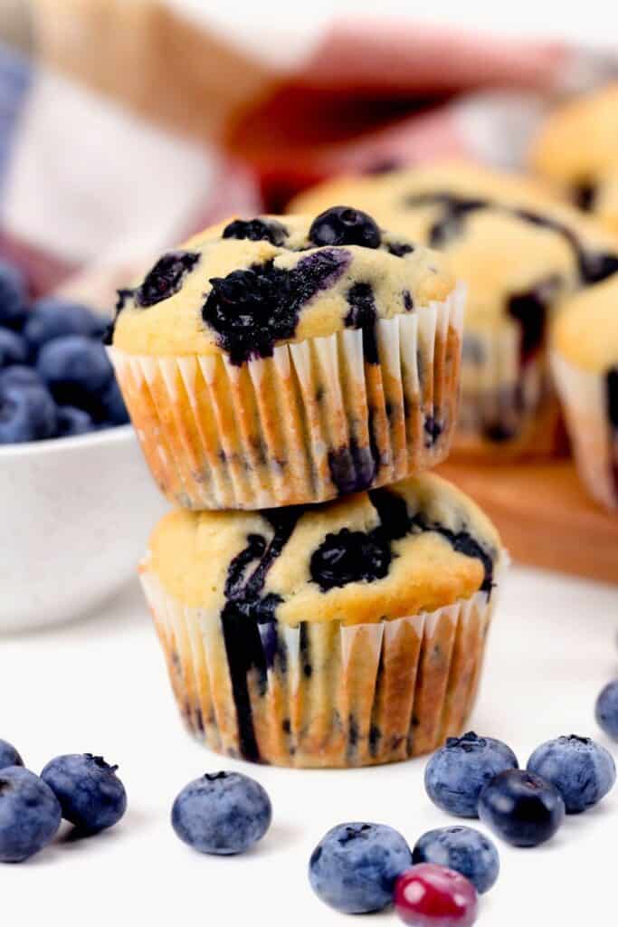 Close up of low sugar blueberry muffins stacked on a white surface with fresh blueberries scattered around. The golden brown tops are lightly domed with juicy blueberries baked throughout. 