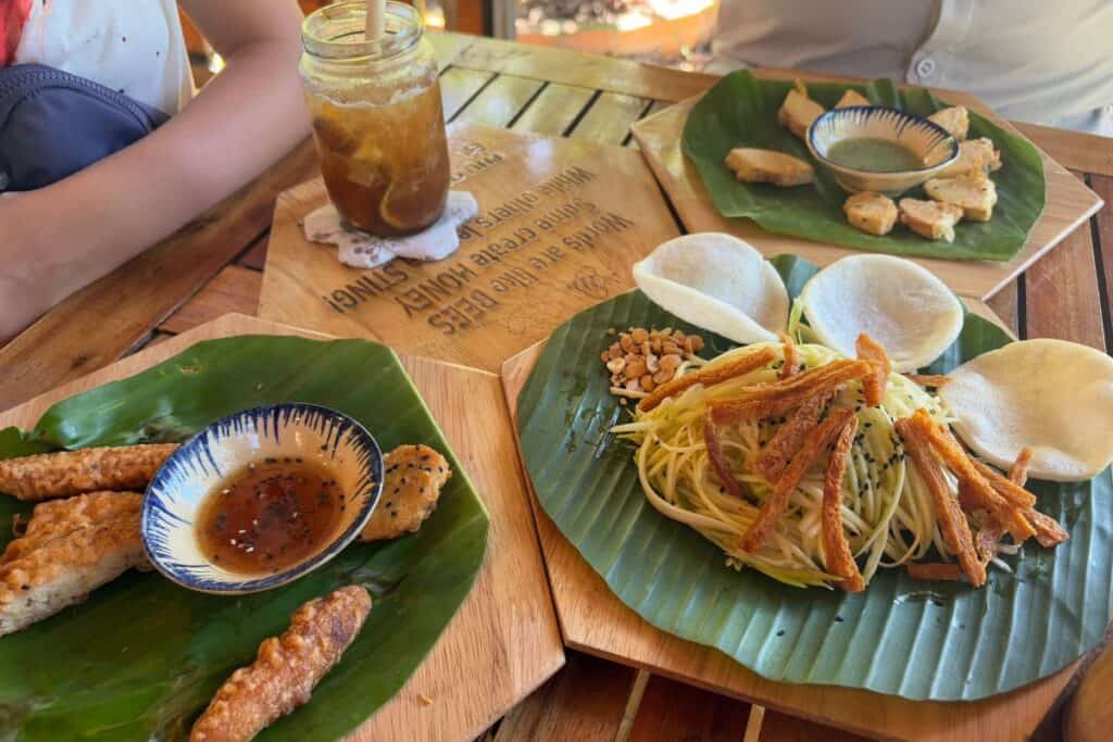 A colorful lunch spread at Phu Quoc Bee Farm featuring a green mango salad with peanuts, sesame seeds, and crispy fried tofu strips, served with rice crackers; fried bread fruit pieces with dipping sauce; and grilled Bee Farm cakes with green sauce, all presented on banana leaves atop wooden platters. An iced herbal tea with honey in a mason jar beside it. 