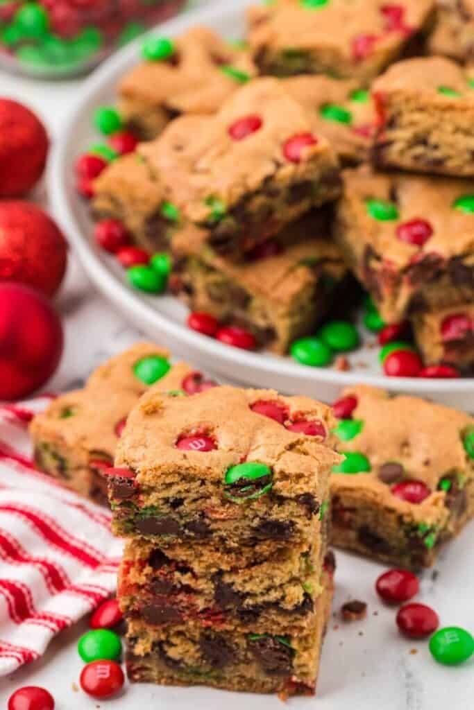 Thick and chewy Christmas cookie bars are loaded with red and green holiday M&M’s and rich chocolate chips, stacked high on a white surface.