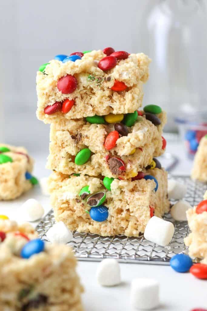 Stacked M and M Rice Krispie treats with colorful candy pieces and small marshmallows visible throughout the soft cereal squares. The treats sit on a wire rack with extra candies scattered around making them fun no bake treats for teens.