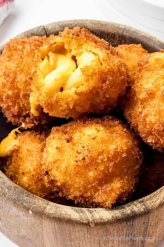 A wooden bowl filled with crispy, golden-brown mac and cheese bites, with one bite split open to reveal creamy, cheesy macaroni inside. Perfectly fried for a crunchy, gooey snack.