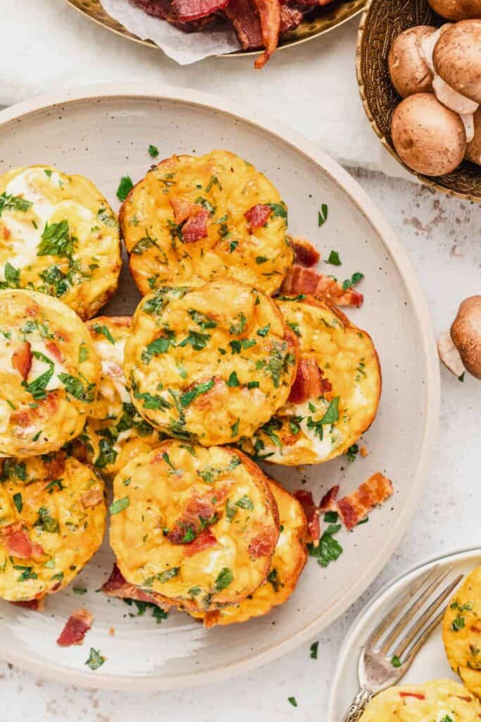 Plate of mini egg muffins with melted cheese bacon and chopped herbs served as a make ahead breakfast muffins option. Several muffins are stacked on a plate with extra bacon pieces and fresh mushrooms visible nearby.
