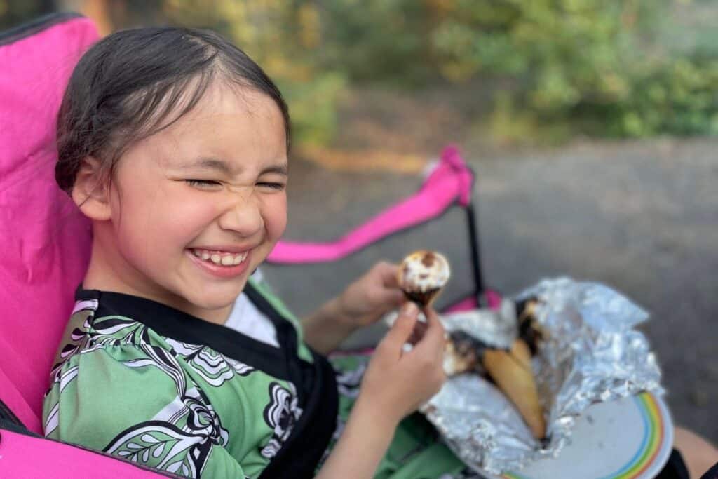 Young girl smiling, sitting in camp chair & eating a s'mores cone.