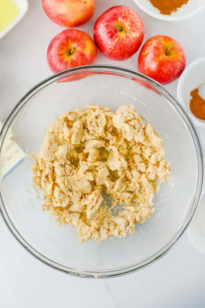 Overhead process shot of crumbly crust dough mixed in a large glass bowl while making apple pie bars. Fresh red apples and bowls of cinnamon and spices surround the dough on the counter.