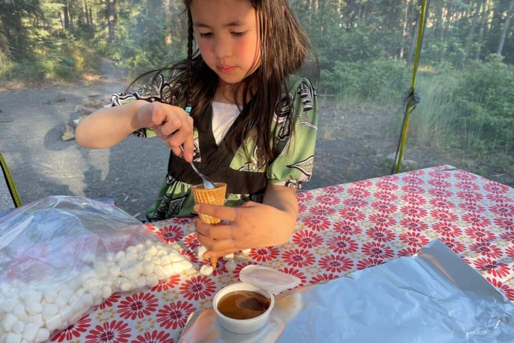 Young girl sitting at a picnic table inside a tent at a campsite, carefully spooning marshmallow topping into an ice cream cone. A bag of mini marshmallows and a small cup of chocolate or caramel sauce sit on the red floral tablecloth, with a wooded campground visible in the background.