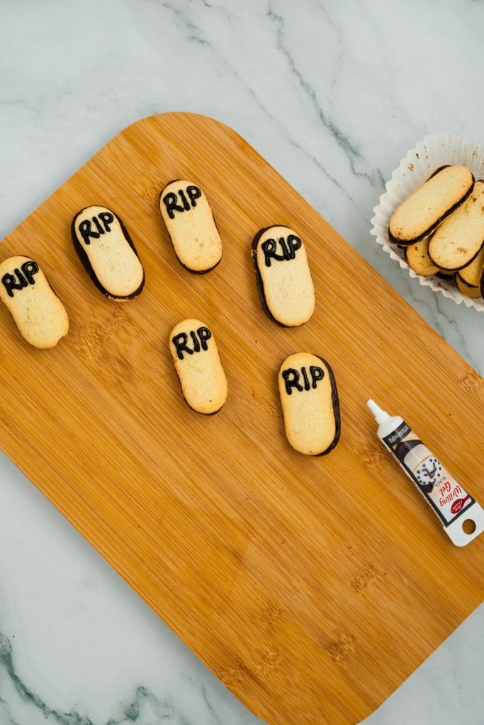 Oval-shaped Milano cookies decorated with “RIP” written in black gel icing are arranged on a wooden cutting board, resembling tombstones for Halloween dirt and worms desserts. A tube of black writing gel and a paper liner filled with plain cookies are nearby.