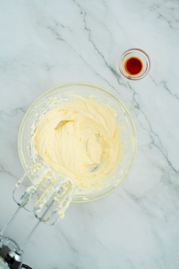 Creamy blended mixture of butter, cream cheese, and powdered sugar in a glass bowl, with an electric hand mixer in the foreground and a small glass bowl of vanilla extract nearby, ready to be added.