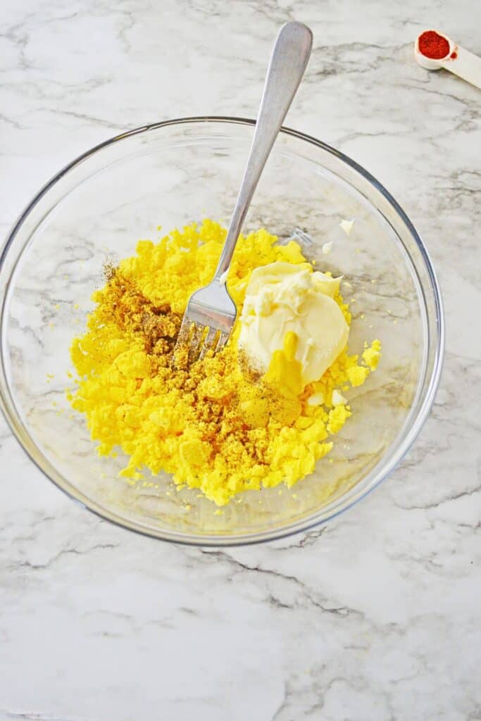 Mashed egg yolks, mayonnaise, mustard, and seasonings sit in a clear mixing bowl with a fork. The ingredients are ready to be mixed into a creamy filling.