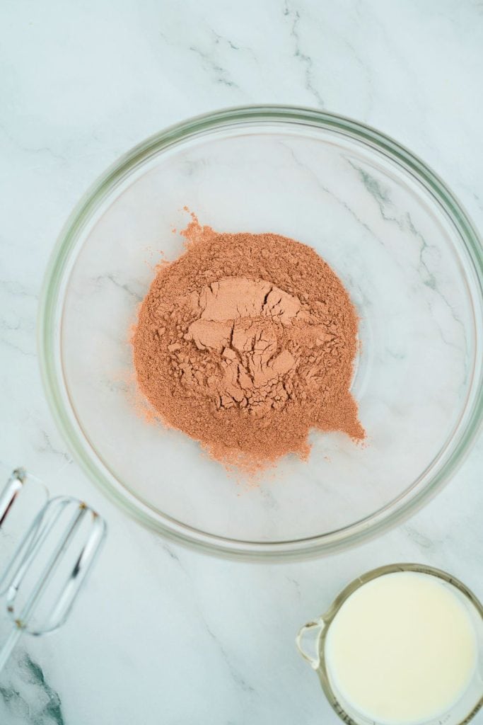 Glass mixing bowl filled with dry chocolate pudding mix on a marble countertop, with a glass measuring cup of milk and electric mixer beaters nearby, ready for preparation.