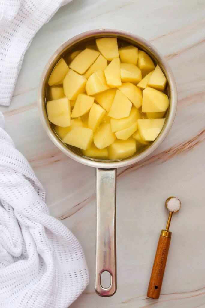 An overhead view of a  medium sized pot filled with cubed yellow potatoes and water on a marble counter top. A tsp of salt is beside the pot. 
