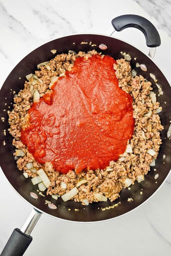 Tomato pasta sauce poured over browned Italian sausage with softened onion and garlic in a large skillet. The meat sauce is ready to be mixed together for the one pan lasagna.