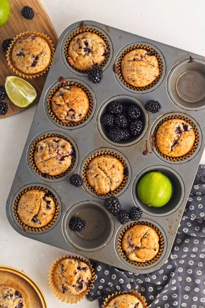 Maple blackberry lime muffins baked in a muffin tin with golden tops and bursts of dark blackberries scattered throughout. Fresh blackberries and whole limes surround the muffins.