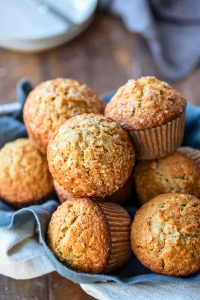 Basket of maple brown sugar oatmeal muffins nestled in a cloth napkin, with golden domed tops and a sprinkle of coarse sugar for crunch. The muffins have a hearty texture and warm, cozy appearance, perfect for a comforting breakfast or snack.