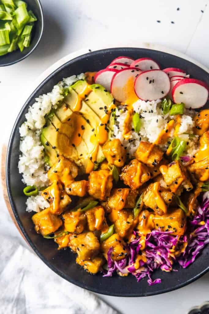 Maple glazed tofu bowl with crispy tofu cubes drizzled in chipotle sauce served over white rice with sliced avocado, radishes, shredded purple cabbage, and green onions. Black sesame seeds and a creamy orange sauce top the colorful tofu bowl.