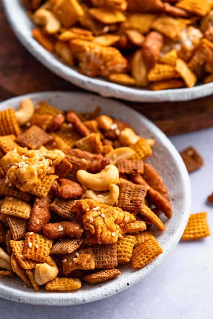 Bowl of maple miso Chex mix made with Chex cereal, nuts, crunchy snack pieces, and sesame coated clusters tossed in a glossy maple miso seasoning. The sweet and savory snack mix is shown close up with a second bowl in the background, highlighting its crunchy texture and variety of ingredients.