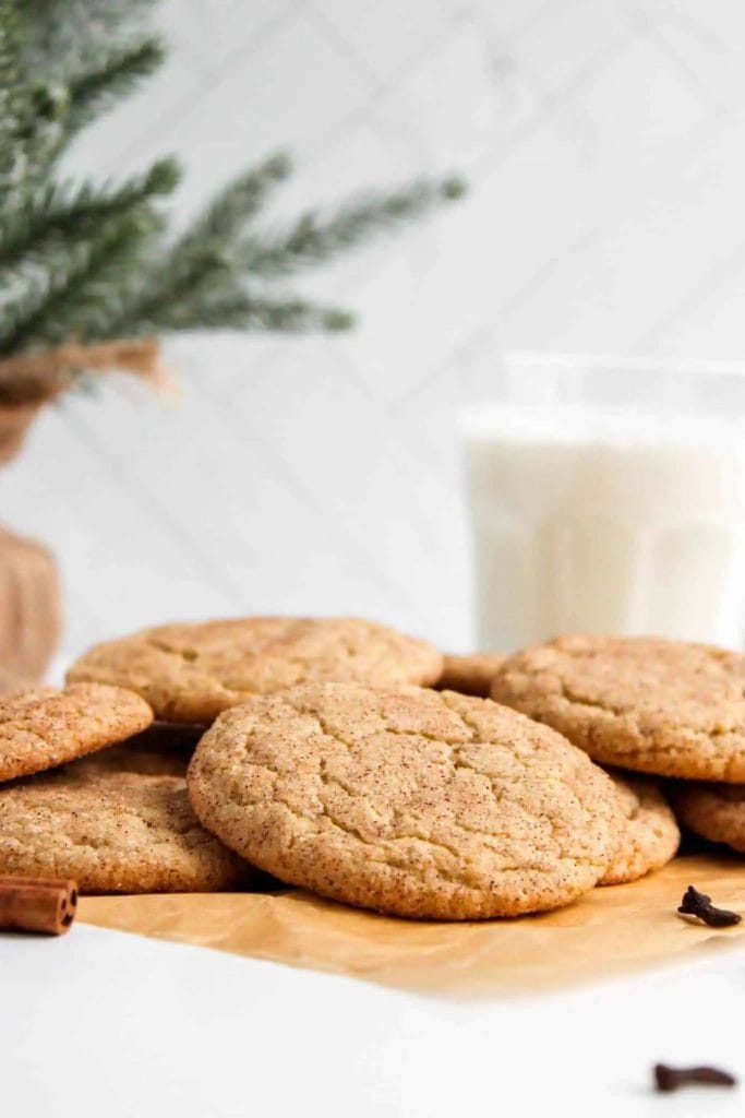 A batch of golden-brown snickerdoodle cookies dusted with cinnamon sugar, arranged on parchment paper. In the background, there's a glass of milk and a sprig of pine, creating a cozy holiday vibe.