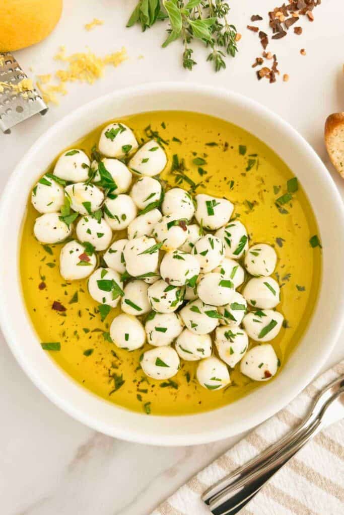 Marinated mozzarella balls are submerged in golden olive oil with chopped fresh herbs, black pepper, and red pepper flakes in a white serving bowl. Fresh herbs, lemon zest, and seasonings surround the bowl.