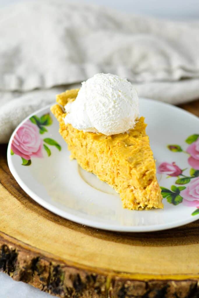 Slice of no bake marshmallow pumpkin pie with a creamy orange filling and golden crust topped with a scoop of whipped topping. The pie is served on a floral plate.