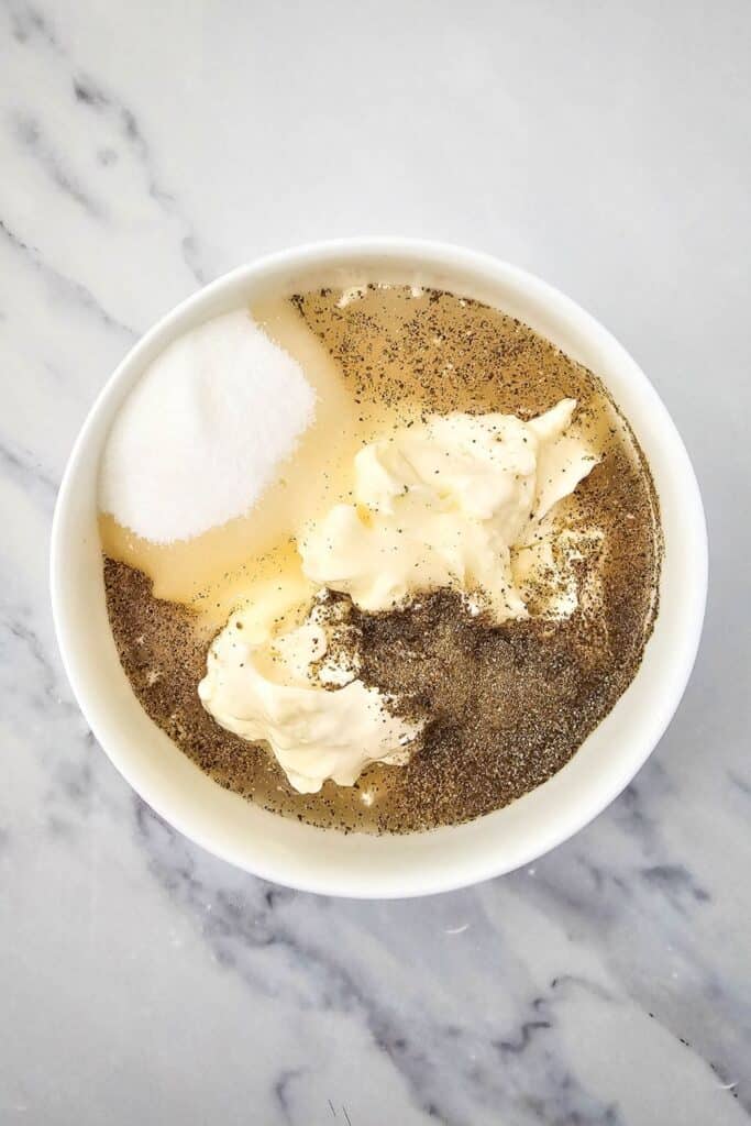 Bowl containing ingredients for a creamy dressing, including mayonnaise, white sugar, black pepper, and white vinegar, set on a marble countertop before mixing