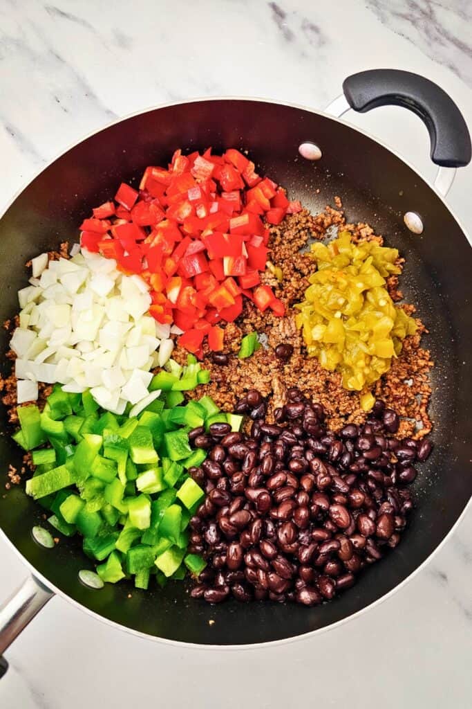 Large skillet filled with cooked seasoned ground beef, topped with colorful ingredients including chopped red bell peppers, green bell peppers, onions, black beans, and green chilies. This vibrant mixture is being prepped for a flavorful sheet pan Tex Mex dinner. 