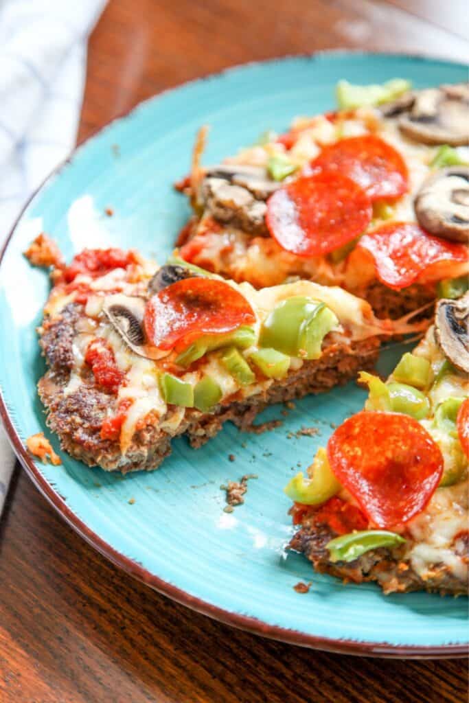 Slice of low carb pizza with a browned meat crust topped with melted cheese, pepperoni slices, green bell peppers, and mushrooms on a blue plate.