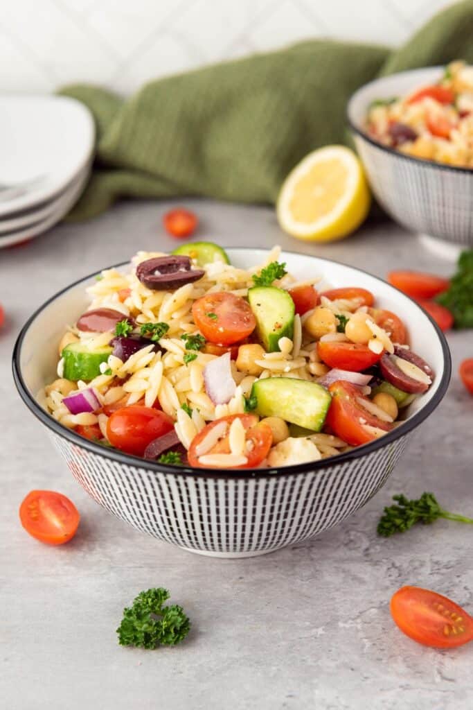 A patterned bowl filled with Mediterranean orzo salad made with orzo pasta, cherry tomatoes, cucumber, chickpeas, Kalamata olives, red onion, feta cheese, and fresh parsley, with lemon in the background.