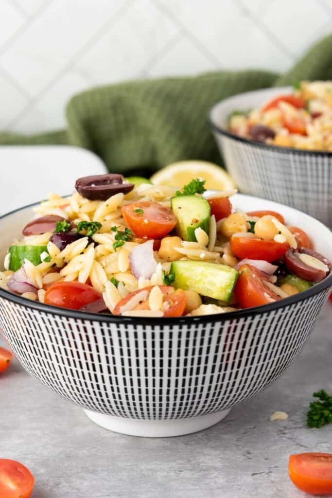 A vibrant Mediterranean orzo salad in a patterned bowl, filled with cherry tomatoes, cucumber, red onion, chickpeas, Kalamata olives, and fresh parsley. A lemon wedge and second bowl of salad are blurred in the background.