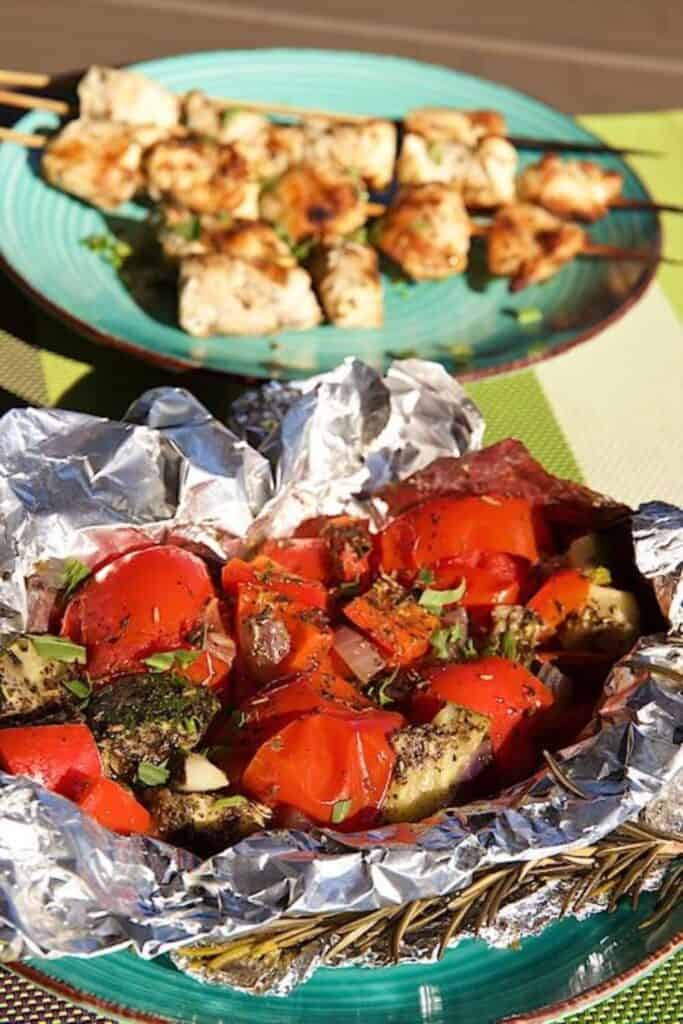 Mediterranean herbed tomatoes, zucchini and peppers in foil packs beside a plate of chicken skewers. 