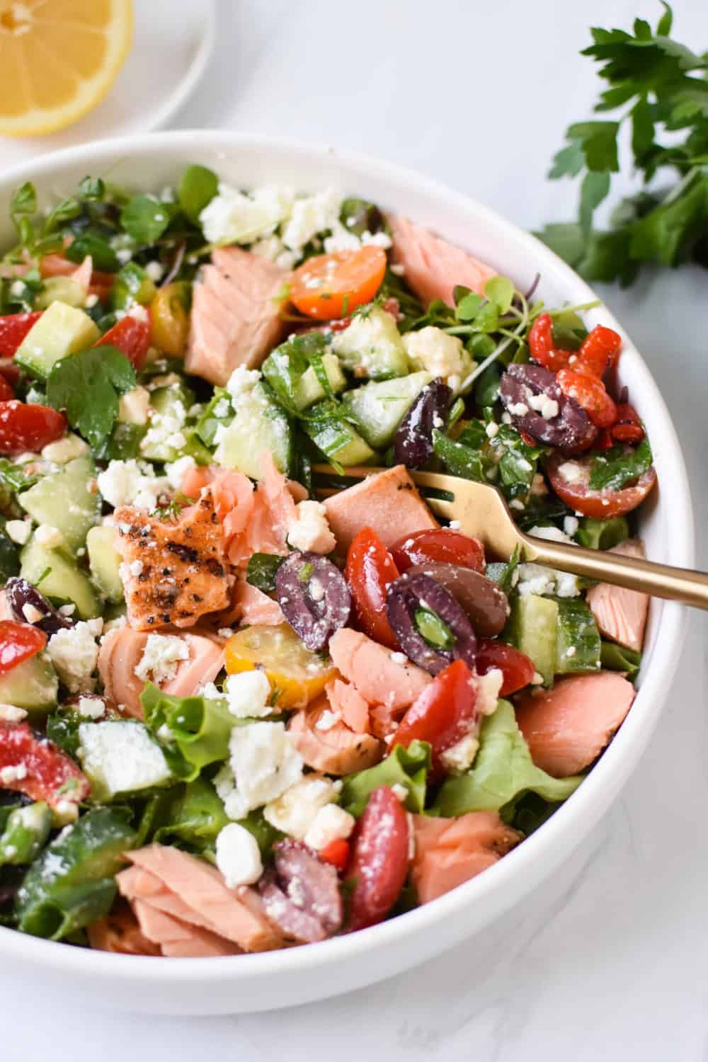Mediterranean salmon bowl with flaky grilled salmon, cucumber, cherry tomatoes, Kalamata olives, crumbled feta, and mixed greens tossed together in a white serving bowl. Fresh herbs and a lemon wedge accompany this colorful dish, making it a healthy option for a last minute meal.