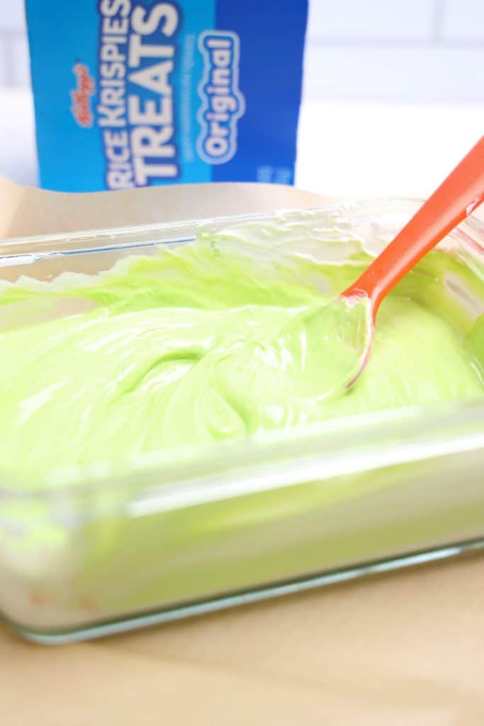 Process shot of a glass dish filled with smooth, melted green candy coating being stirred with a red spoon, set on a sheet of parchment paper.