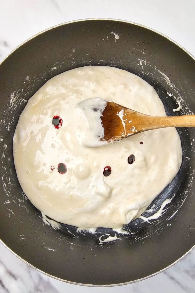 Melted marshmallows in a saucepan with drops of red food coloring, being stirred with a wooden spoon to make Halloween rice krispie treats.