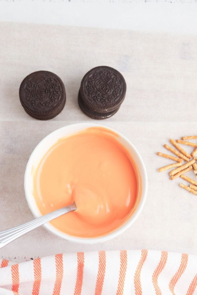 A bowl of melted orange candy with a spoon rests on parchment paper, surrounded by Oreo cookie stacks and scattered pretzel sticks. An orange and white striped towel lines the bottom edge, suggesting a setup for making festive treats.